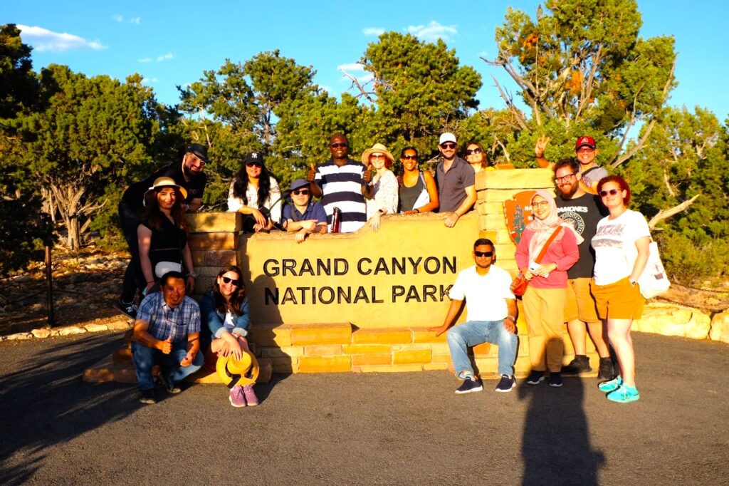 The scholars pose around a sign marking the Grand Canyon National Park, squinting against the late afternoon sun