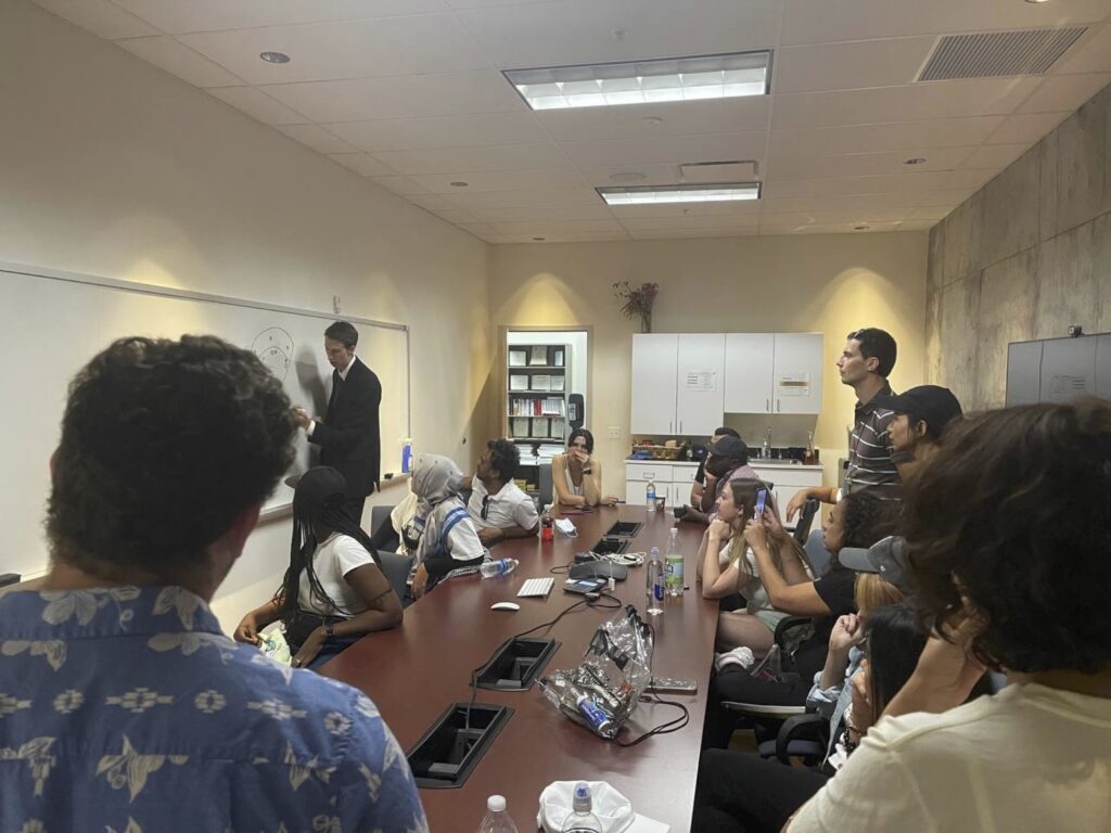 The scholars surround a conference table while watching Andrew Lind diagram baseball on a whiteboard