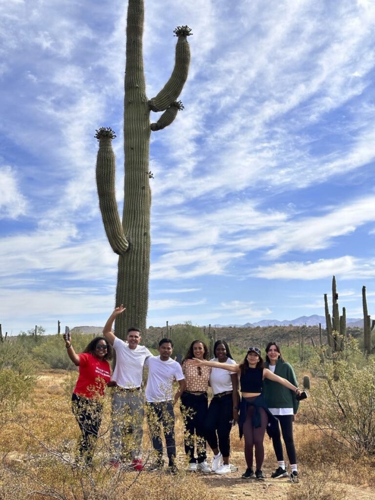 A group of scholars poses under a tall saguaro cactus; the sky and desert stretch out in all directions