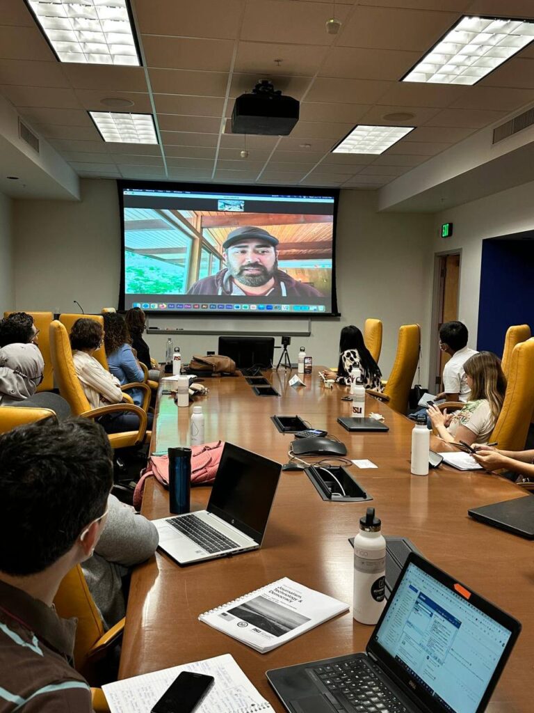 The scholars sit around the conference table in Room 444, with Dennis "Fox" Bonilla on the Zoom screen