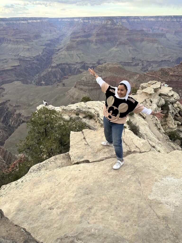 A woman stands in a "ta-da!" pose on a rock overlooking the Grand Canyon