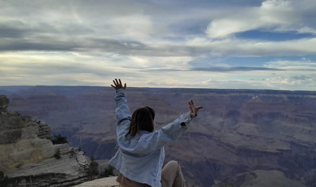 A women sits, arms outstretched as if in benediction, on a rock overhanging the vast expanse of the Grand Canyon