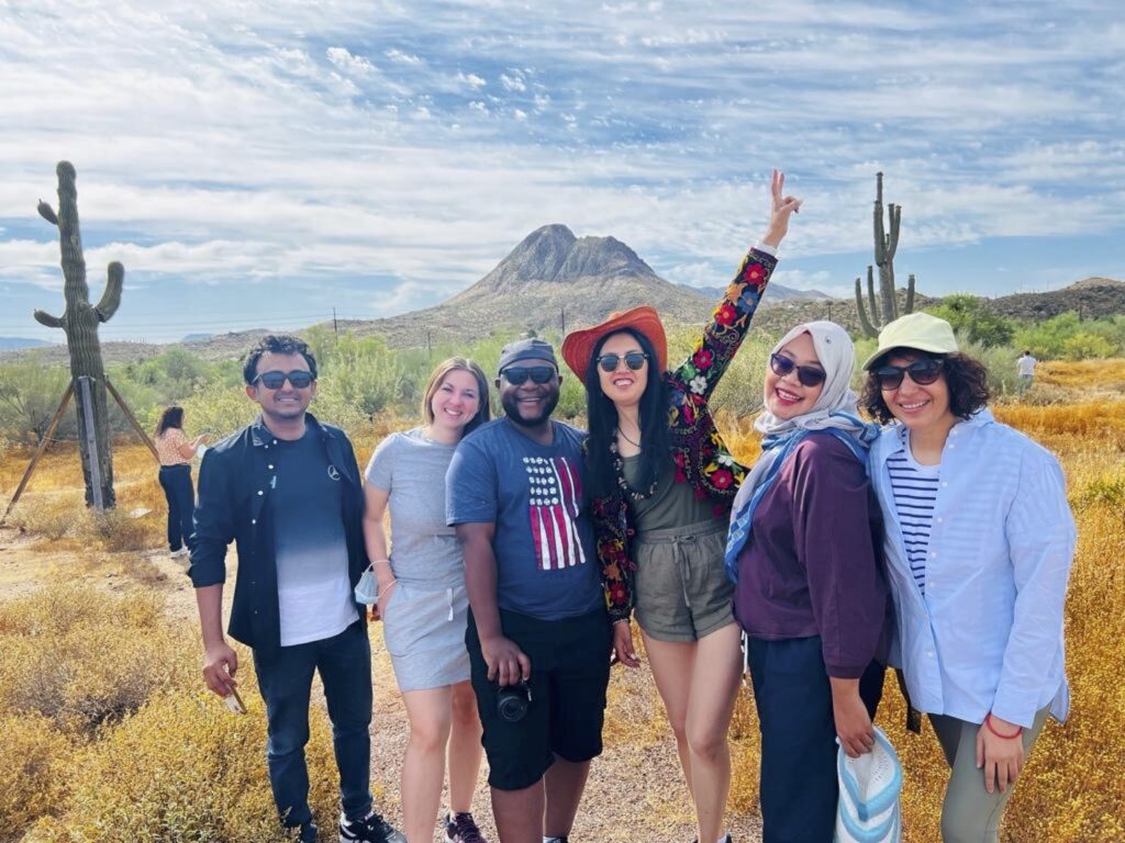 A group of smiling scholars stands in the Arizona desert amid the saguaro cacti