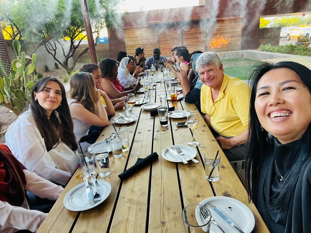 The scholars and Dan Barr sit around an outdoor restaurant table, steam from the misters visible overhead