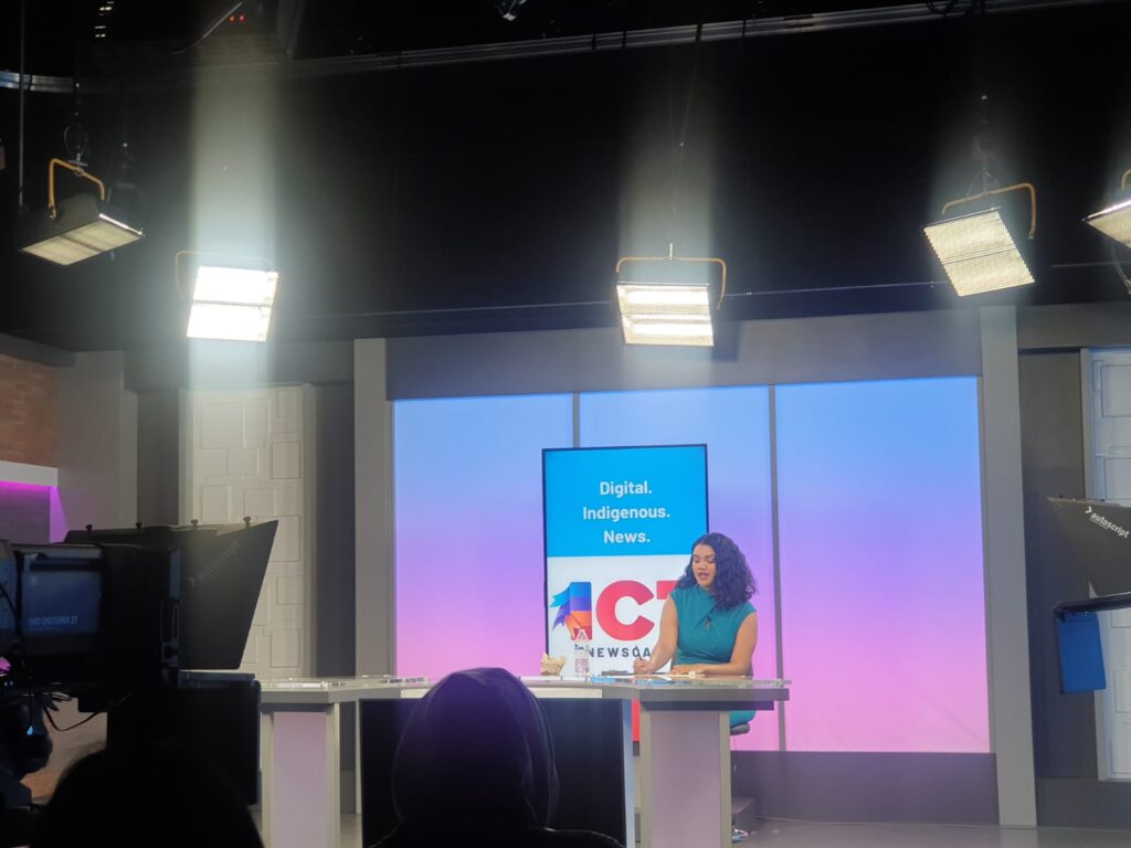 A young woman sits at a news anchor desk in front of the ICT logo