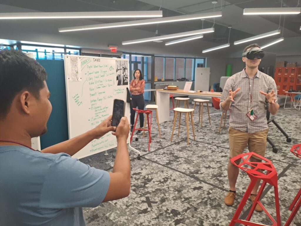 A scholar wearing a VR headset stands in a room, while another records him