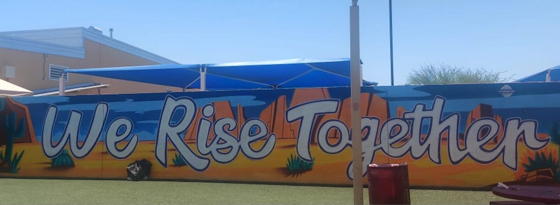 A mural reads "We Rise Together," with desert images and a saguaro cactus