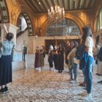 A group of scholars observe a tour guide pointing out architectural details in an ornate Art Deco-era building