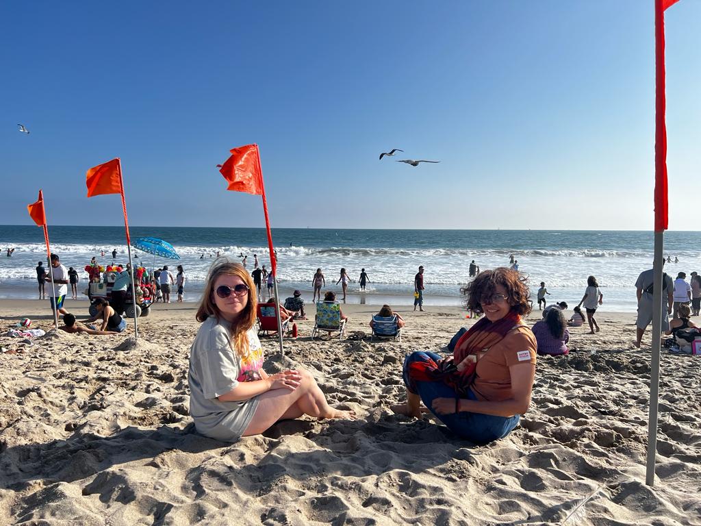 Two women sit on a beach, with waves and seagulls