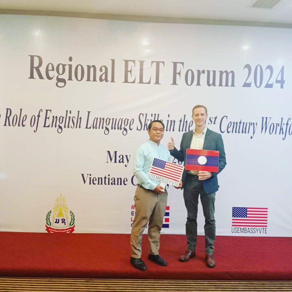 Two gentlemen standing front of a sign proclaiming "Regional ELT Forum 2024." One is holding a US Flag, the other a Laotian flag