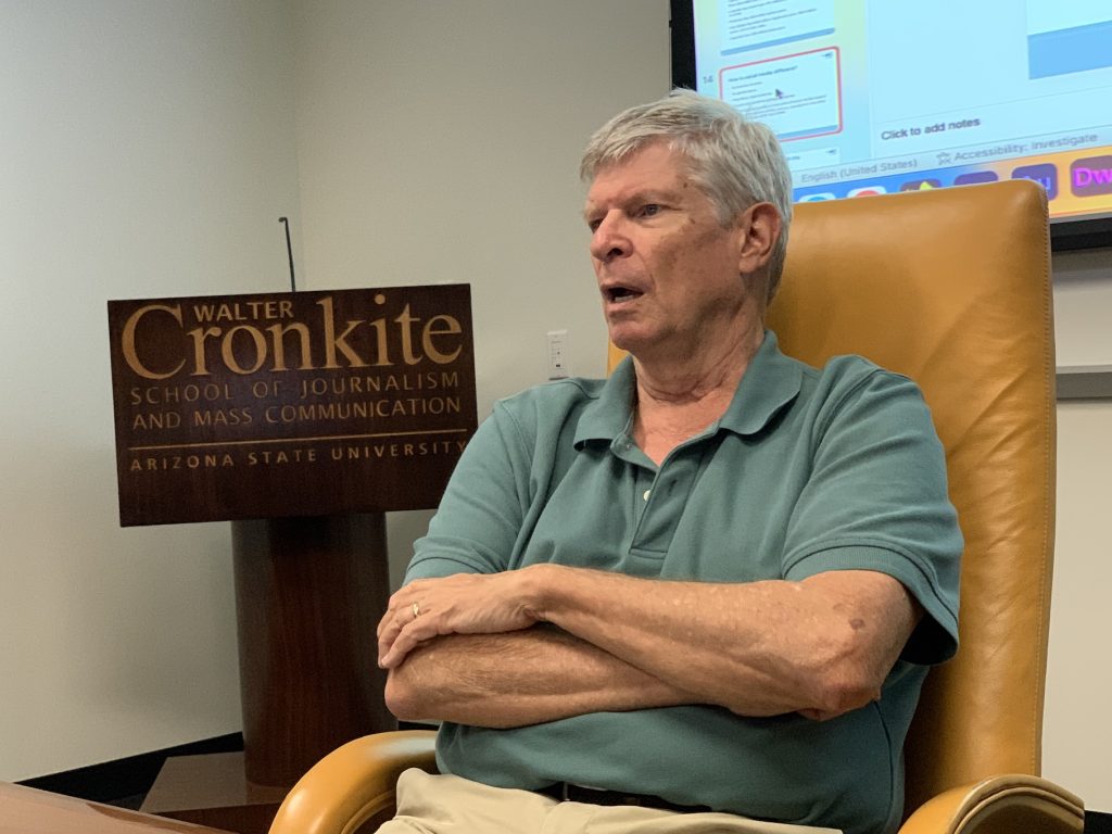 A man sits with folded arms in a yellow high-backed conference chair, in front of a podium branded by ASU's Cronkite School