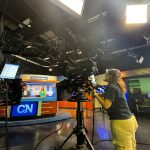 The Cronkite News studio during a newscast, with student anchors at the desk and another student operating the camera in the foreground