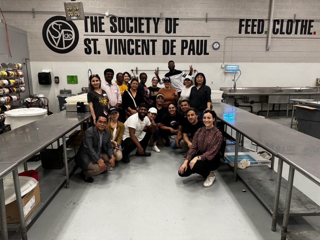 The scholars and Kate Devine pose in a clean industrial kitchen under the logo of the Society of St. Vincent De Paul