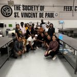 The scholars and Kate Devine pose in a clean industrial kitchen under the logo of the Society of St. Vincent De Paul