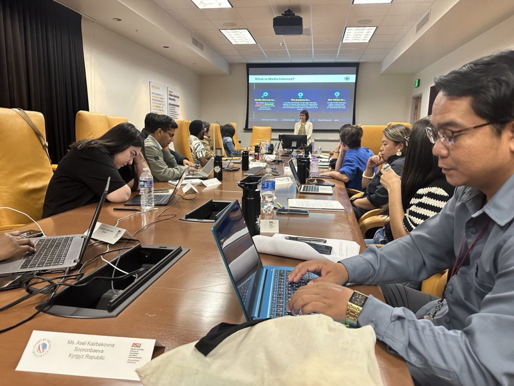 The scholars take careful notes and use tools on their laptops while Dr. Kristy Roschke addresses them in Cronkite room 444