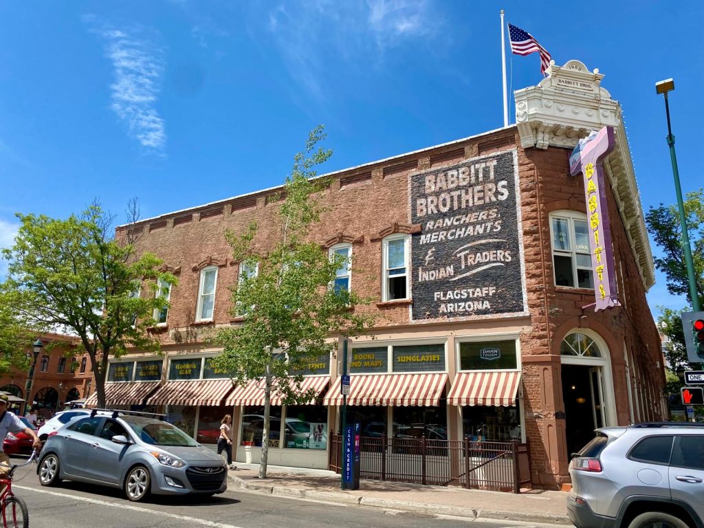 An old-fashioned brick building flying an American flag bears a painted sign identifying it as "Babbitt Brothers - Ranchers, Merchants, & Indian Traders - Flagstaff, Arizona"