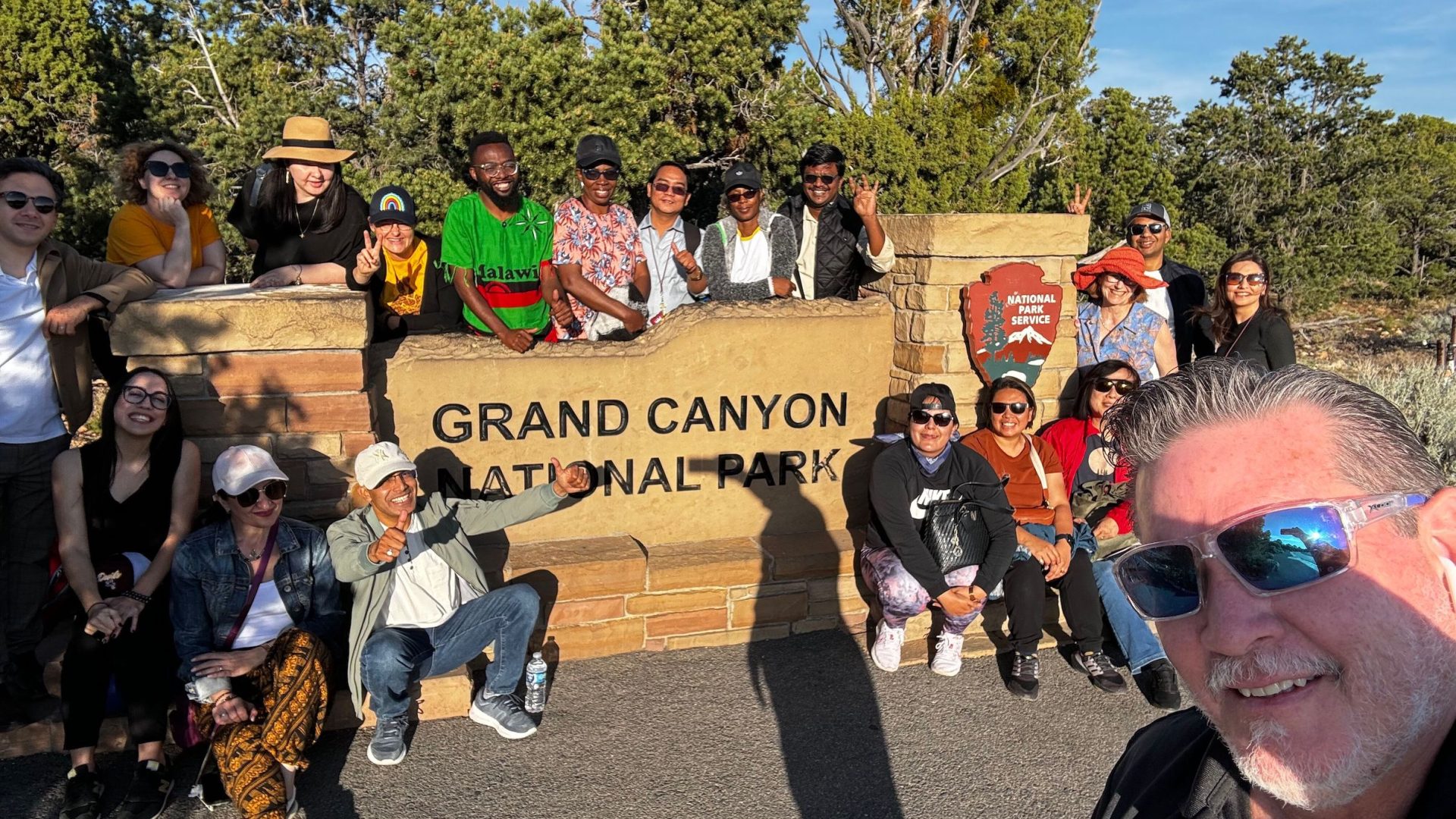 The scholars (with bus driver Ronnie in the foreground) pose around the stone sign marking Grand Canyon National Park