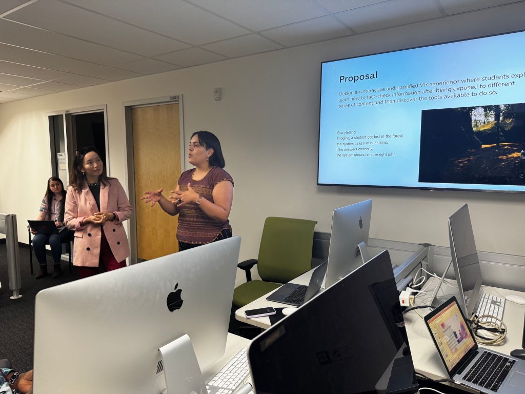 Two women speak animatedly in front of a slide on a screen behind them, surrounded by computer equipment