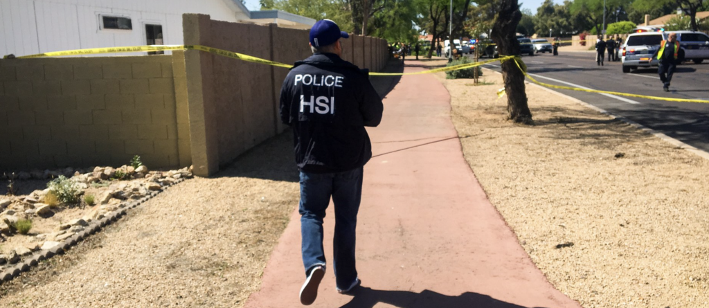 A Homeland Security officer, back to the camera, strides along the sidewalk in a residential neighborhood. Police cars, officers, and yellow crime scene tape complete the picture